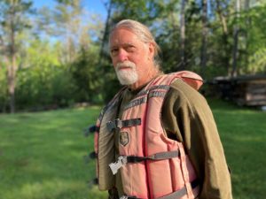 A man with a white beard, wearing a faded life jacket, looks into gentle sunlight