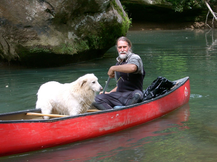 a man, Russ Miller, paddles a red canoe with a white dog and trashbags in the back