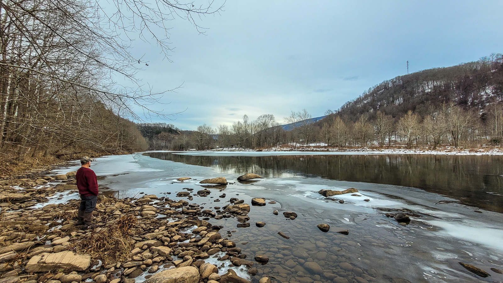 a man in a dark red sweater and camo hat gazes across a river at trees. It is winter, there is ice on the edges of the water and the trees are bare. It is beautiful. 