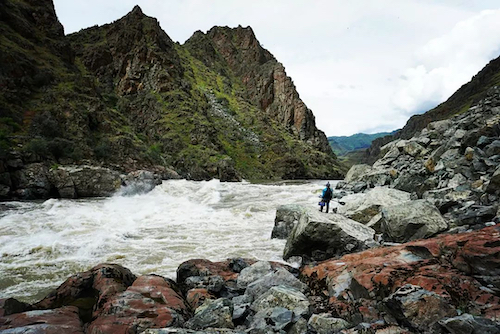 A person stands amid large rocks and boulders, looking at a river rapid