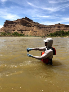 A person stands waist deep in a river that flows through desert landscape, pouring water from one container to another