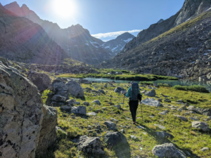 a hiker with trekking poles walks around boulders into a high alpine landscape