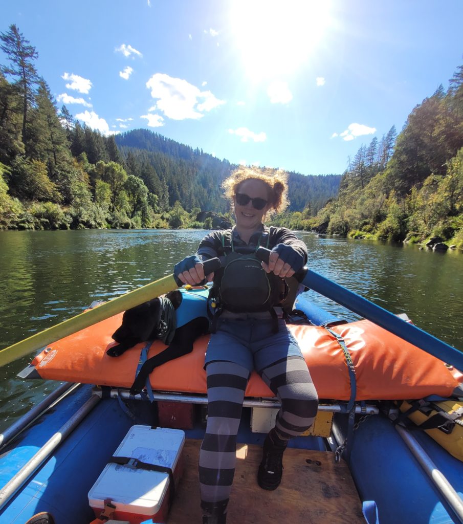 A young woman in sunglasses smiles from the rowing seat of a raft, green mountains and river visible in the sunlight behind her.