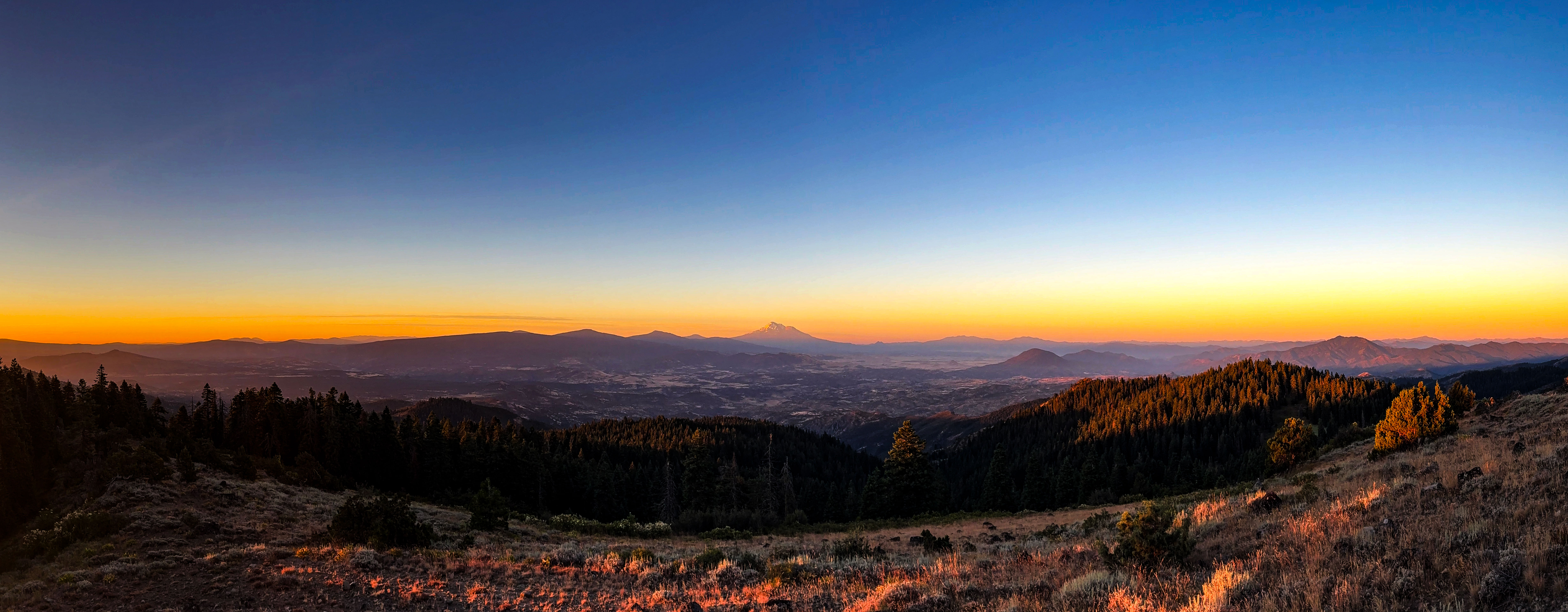 Sunset over forested ridges and grassland seen from Soda Mountain, with a snowcapped peak on the horizon